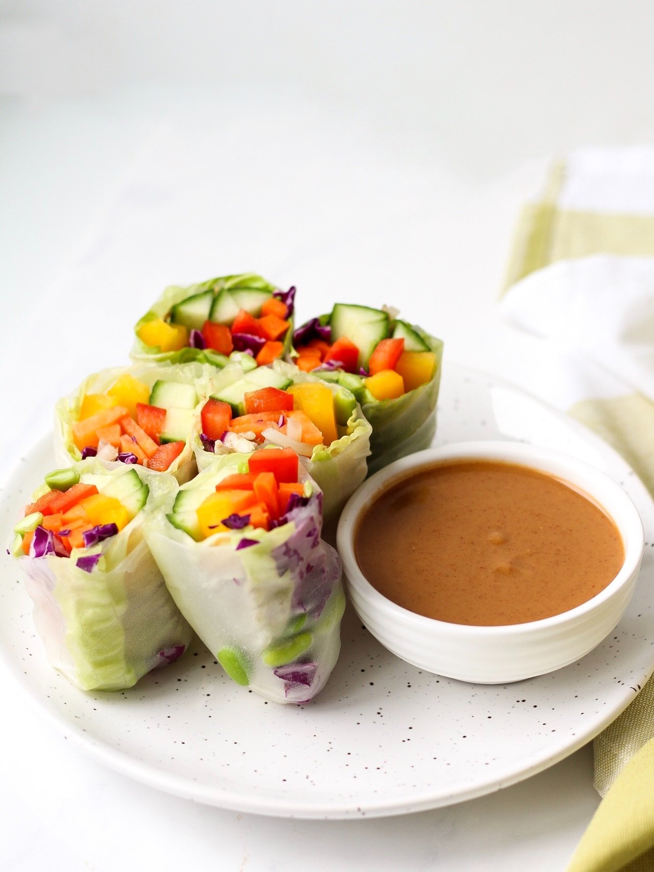 Fresh vegetable spring rolls on a white speckled plate beside a dish of peanut sauce
