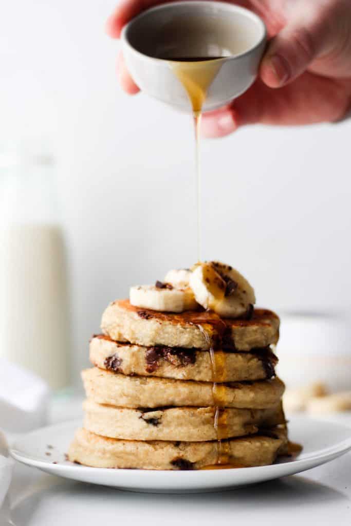 Stack of fluffy chocolate chip pancakes topped with banana. Hand pouring a dish of maple syrup over top
