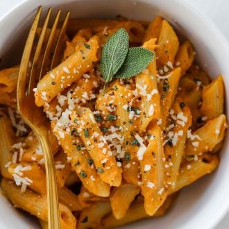 Penne pasta with creamy pumpkin sage pasta sauce garnished with sage leaves and parmesan cheese in a white bowl.