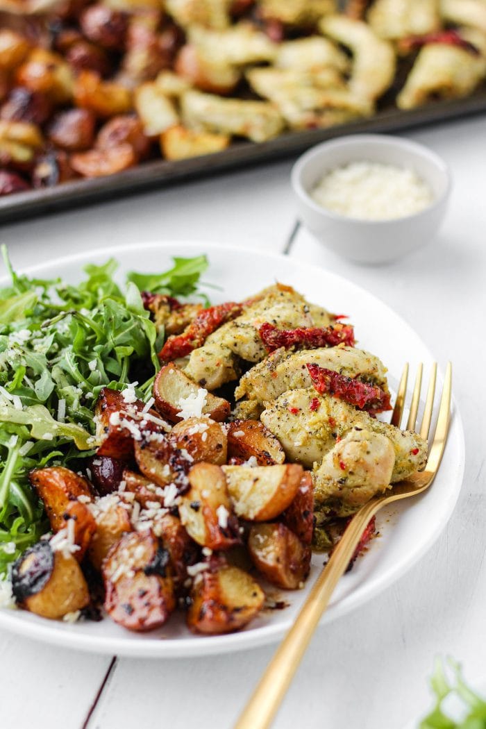 sheet pan pesto chicken and potatoes on a plate with arugula and a pan of chicken and potatoes in the background