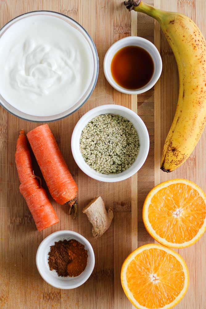 ingredients on a wooden board including yogurt, a banana, a carrot, an orange, ginger root, hemp seeds, and spices