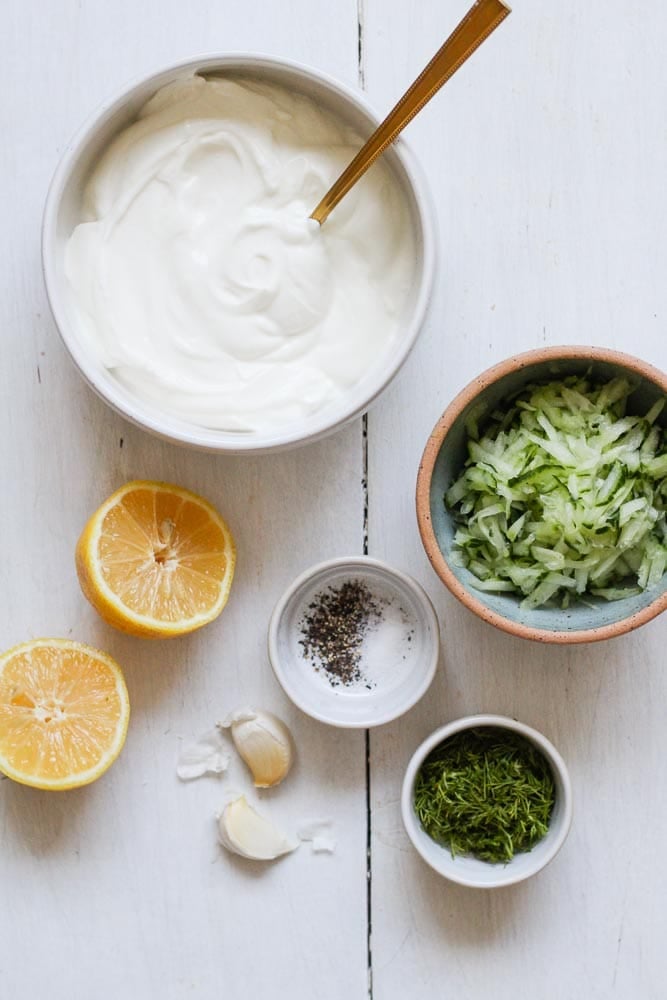 flatlay image of homemade tzatziki ingredients including greek yogurt, lemon, grated cucumber, chopped dill, garlic cloves, salt and pepper