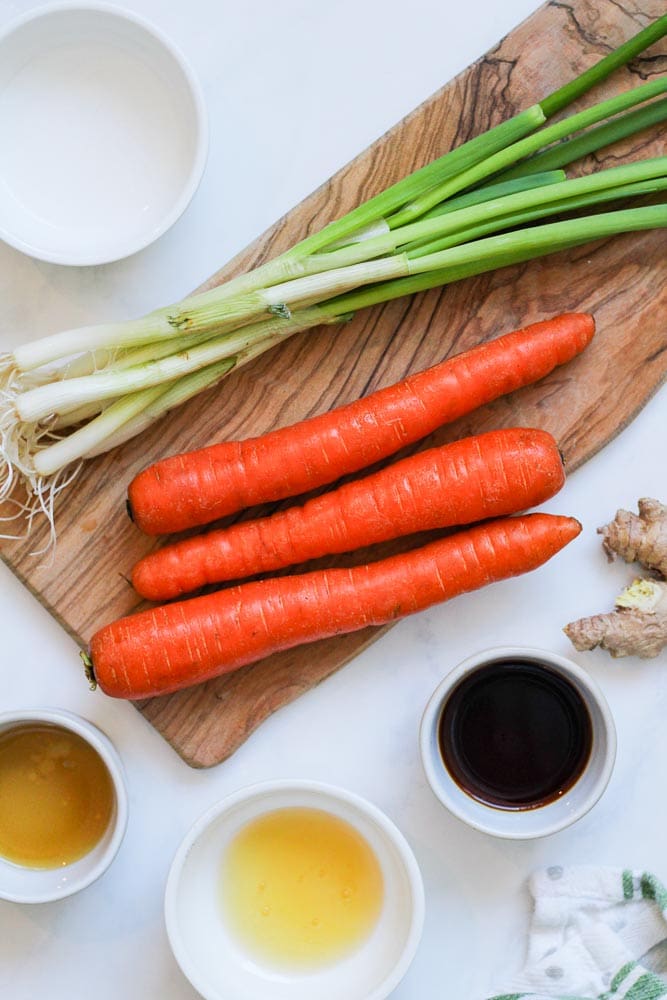 carrots and green onions on a wooden board surrounded by bowls of vinegar, soy sauce, ginger, sesame oil and honey