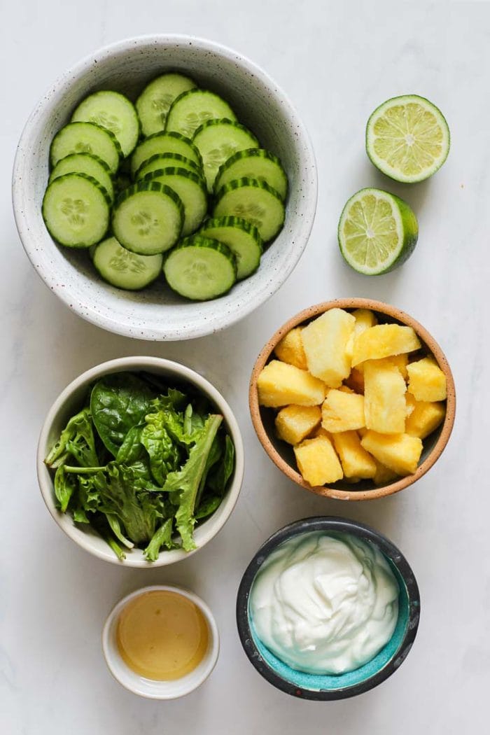 cucumber smoothie ingredients in bowls: sliced cucumbers, frozen pineapple, spring mix greens, greek yogurt, lime, and honey