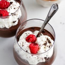 spoon in a glass of blended chocolate chia pudding topped with whipped cream, two raspberries, and chopped chocolate
