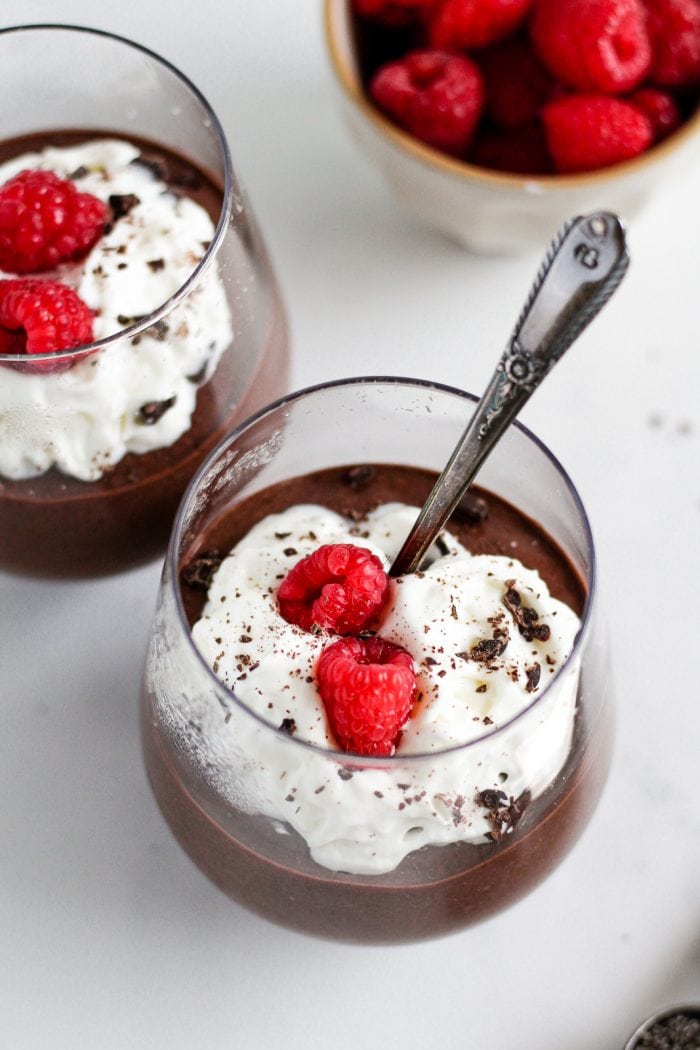 spoon in a glass of blended chocolate chia pudding topped with whipped cream, two raspberries, and chopped chocolate
