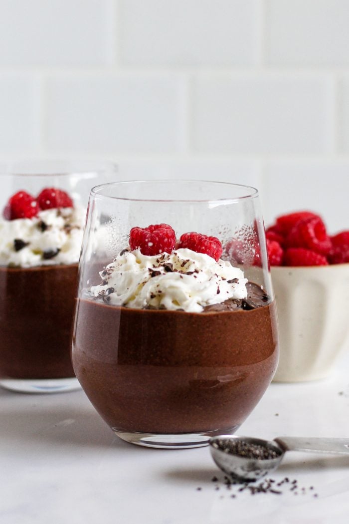 two glasses of blended chocolate chia pudding topped with whipped cream, two raspberries, and chopped chocolate beside a bowl of raspberries