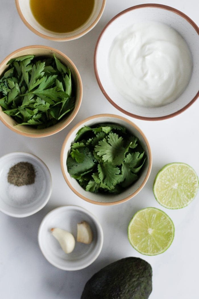 ingredients for greek yogurt avocado dressing including plain greek yogurt, avocado, lime, cilantro, parsley, garlic cloves, salt, pepper, and olive oil