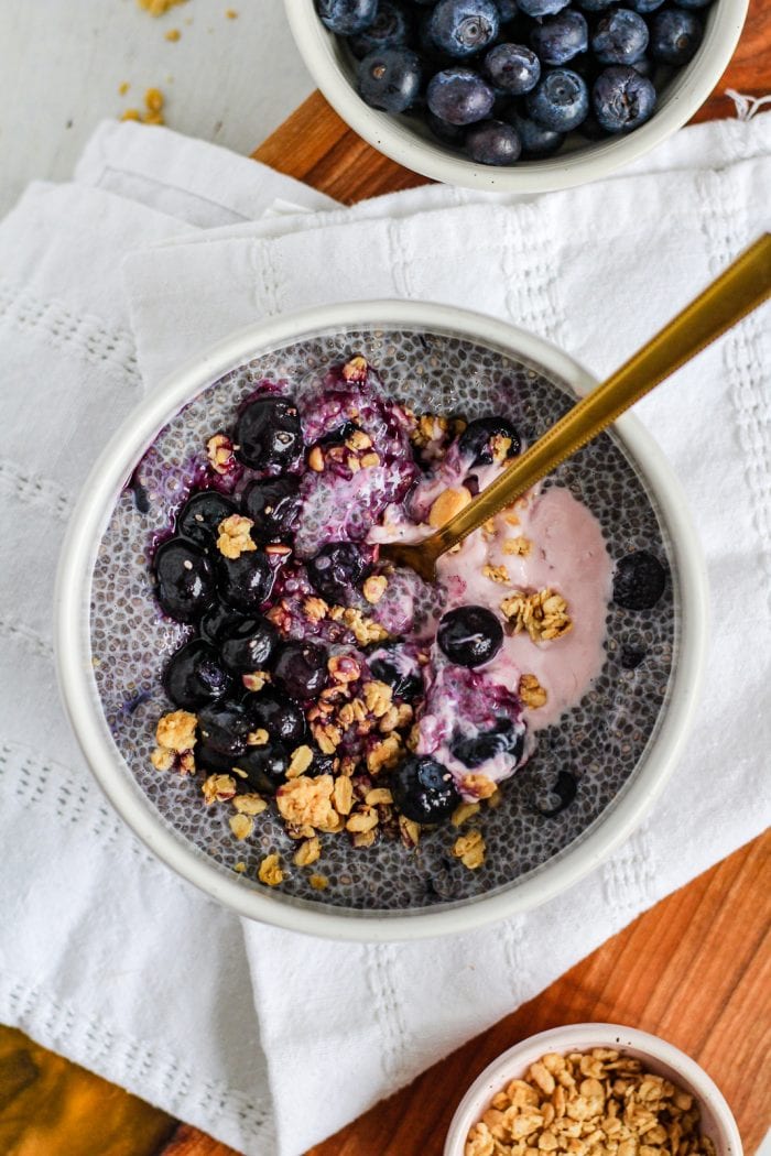 bowl of blueberry chia pudding topped with blueberries, granola, and berry yogurt with a gold spoon in it