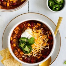 two bowls of beef chili with beans and vegetables topped with plain greek yogurt, shredded cheese, and jalapenos