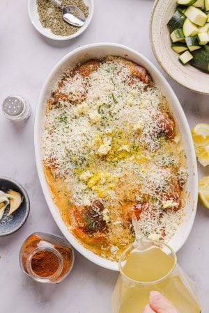 vegetable broth being poured into a casserole dish of chicken thighs covered in rice, spices, garlic, lemon zest and lemon juice