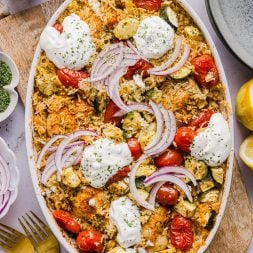 chicken and rice casserole with zucchini, tomatoes, and topped withs sliced red onion and dollops of tzatziki