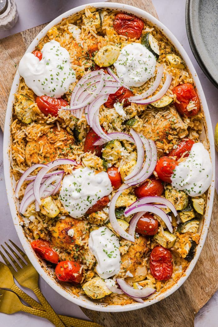 chicken and rice casserole with zucchini, tomatoes, and topped withs sliced red onion and dollops of tzatziki