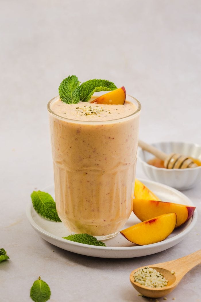 peach smoothie in a glass resting on a plate with peach slices