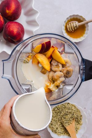 hand pouring a jug of milk into a blender container with peaches, greek yogurt, frozen banana, hemp seeds, vanilla, and honey